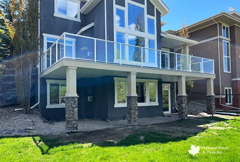 DekSmart vinyl walkout deck with custom Smart Board and stone pillers, and aluminum railings with clear glass inserts.